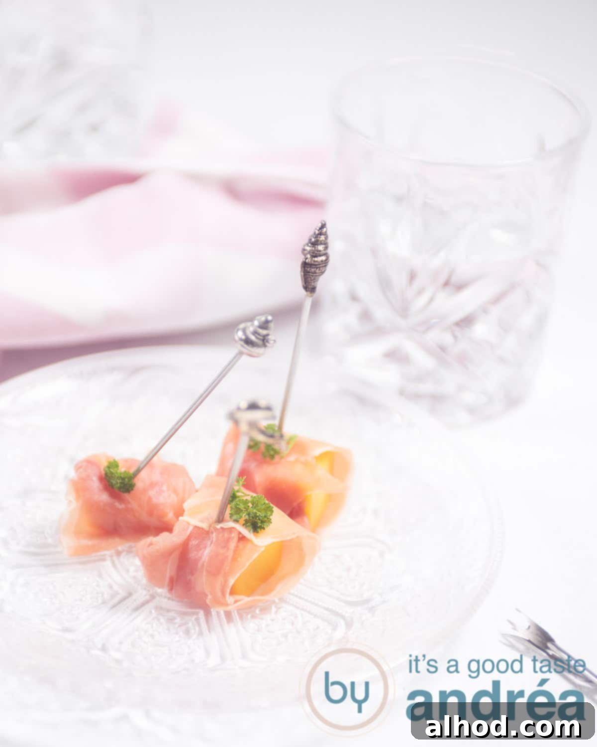 Three prosciutto wrapped mango cubes with aluminum prickers. A pink cloth in the background and a glass to the right