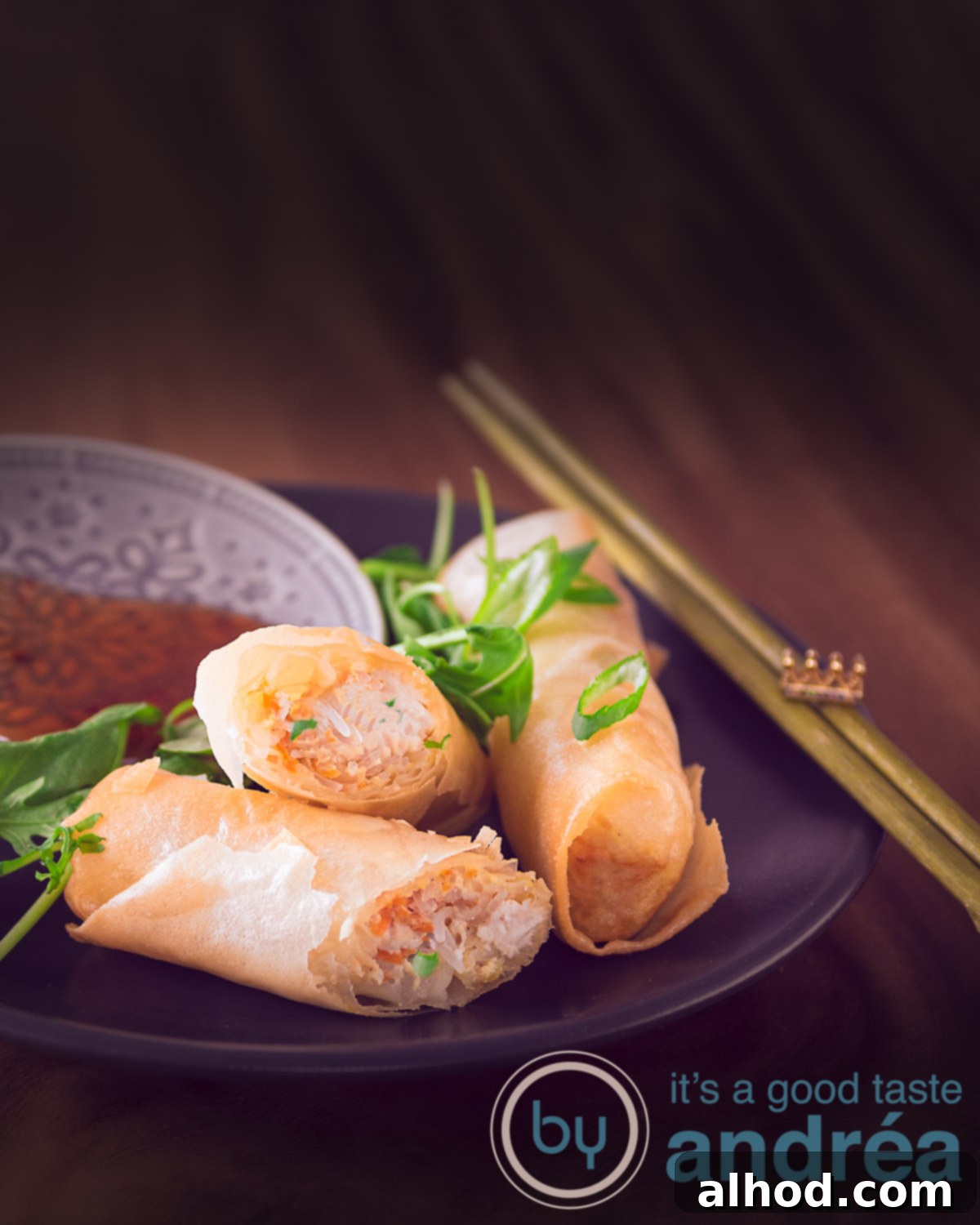 A gray plate with a spring roll and a second one cut in half (showing the contents). A wooden background. To the right two gold chopsticks.