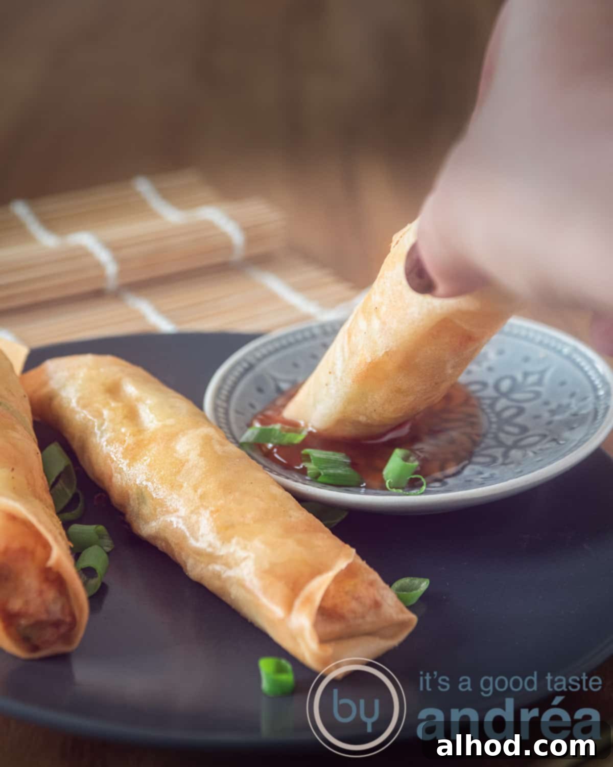 Chicken Lumpia 7 three chicken lumpia, two on a grey plate, one dipped into sauce. In the background a bamboo mat.
