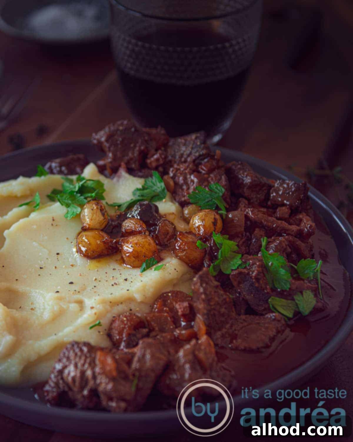 Slow Cooker Beef Bourguignon Without Mushrooms 2 A plate of creamy mashed potatoes, topped with rich slow cooker beef bourguignon and glistening caramelized onions, all set against a rustic dark background.