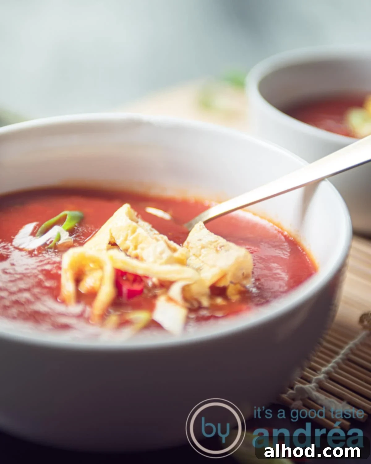 An aerial view showing a bowl of vibrant red Chinese tomato soup with golden egg strips, fresh green spring onion, and a sprinkle of pepper. The bowl sits on a rustic wicker mat, with a golden spoon ready in the foreground, highlighting its inviting and flavorful presentation.