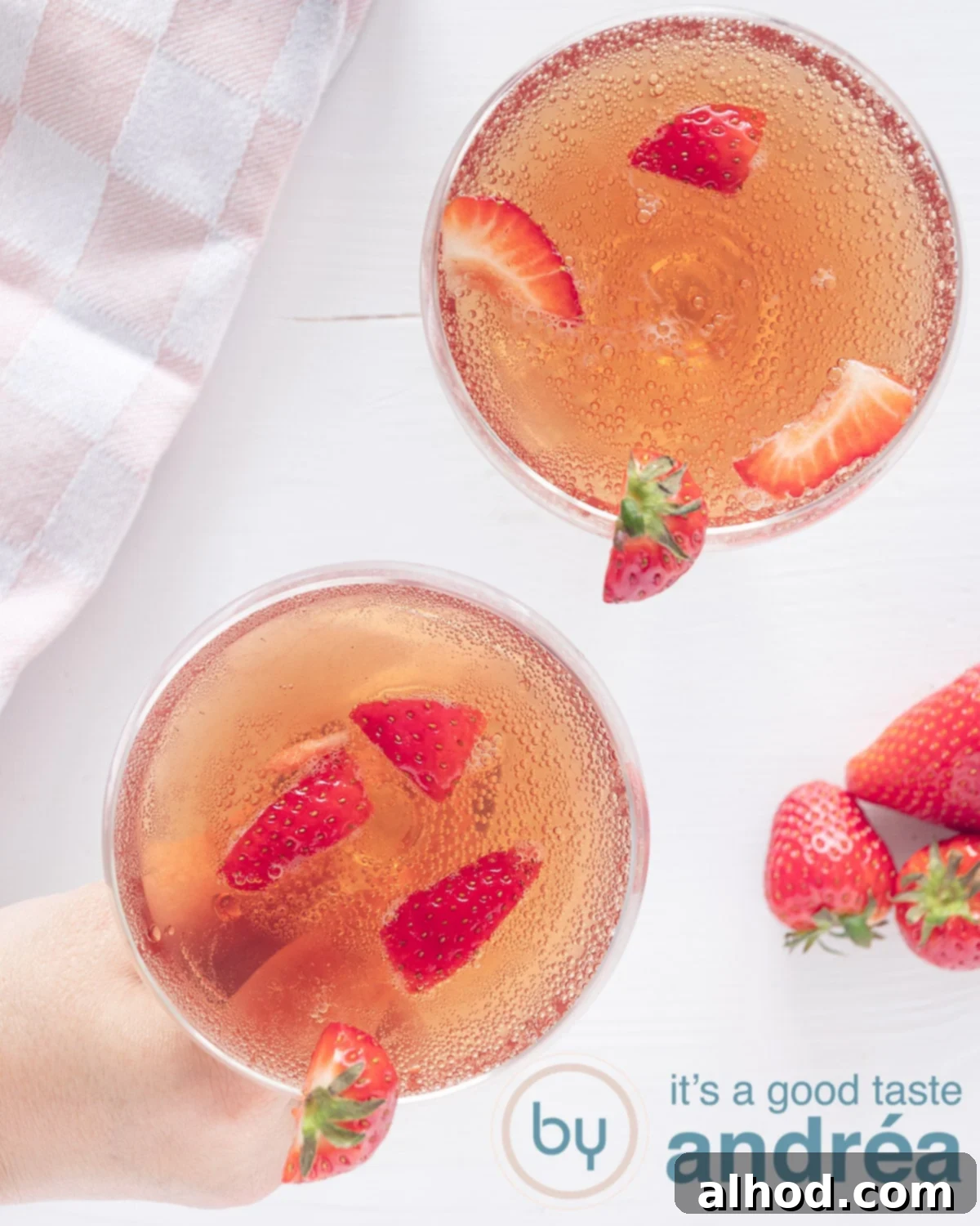 Strawberry Prosecco Fizz 2 A photo from above showing two champagne bowls filled with fresh strawberries and Prosecco on a white background. A hand grabs a glass, ready to enjoy this refreshing sparkling drink.