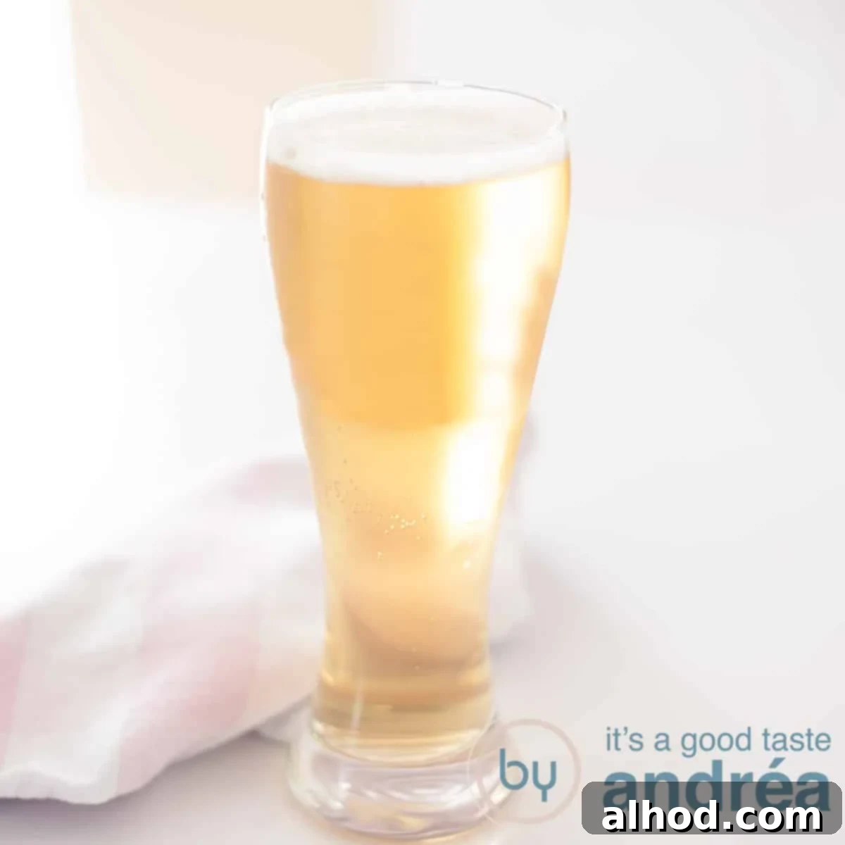 A square photo with a beer cocktail of lager and apple cider on a white surface. A pink white tea towel behind it, suggesting a rustic and refreshing blend.