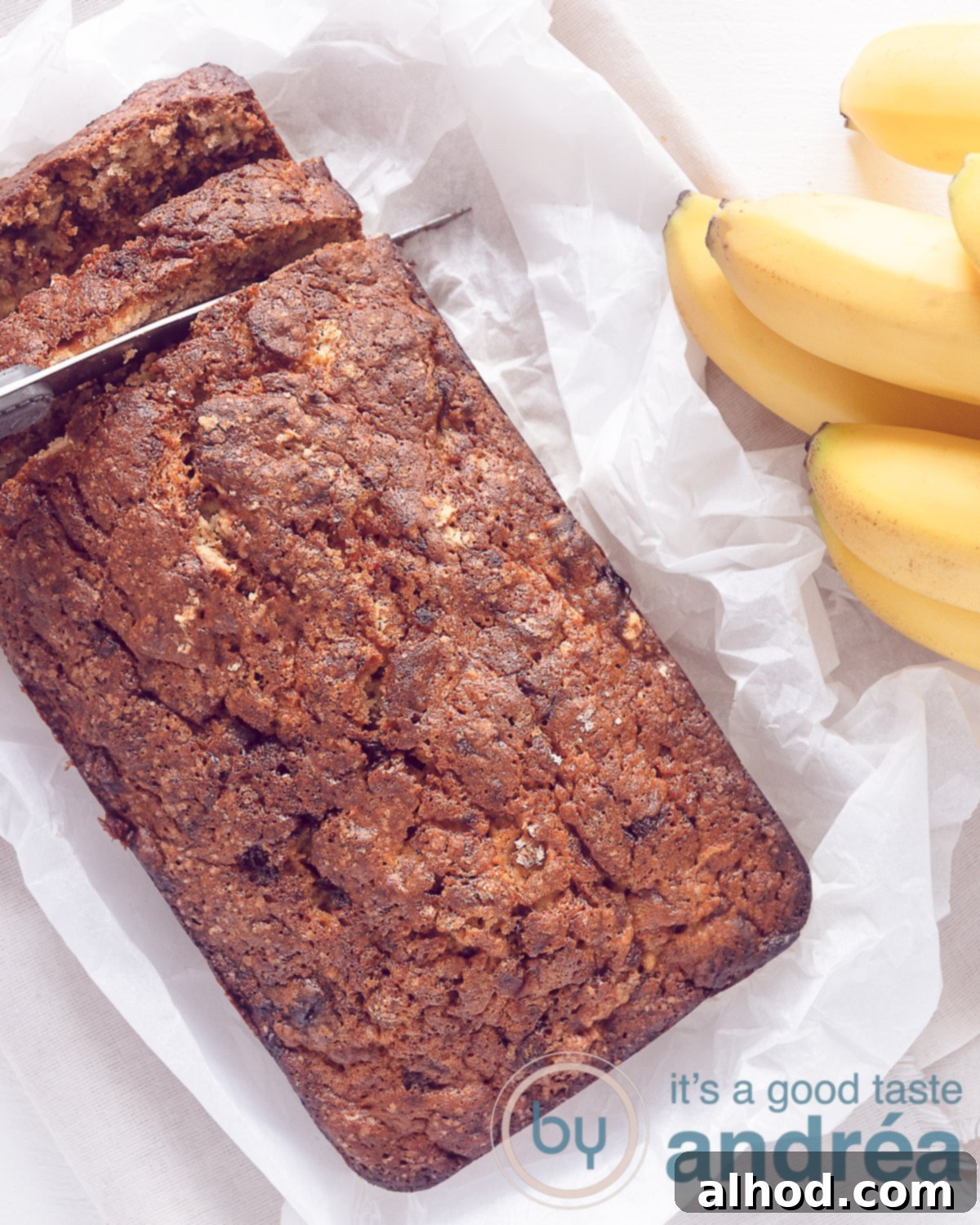 Golden Banana Pound 2 A photo from above of a delicious, moist banana loaf pound cake resting on baking paper, with a knife cutting a slice. A ripe banana bunch is visible on the right, highlighting the key ingredient.