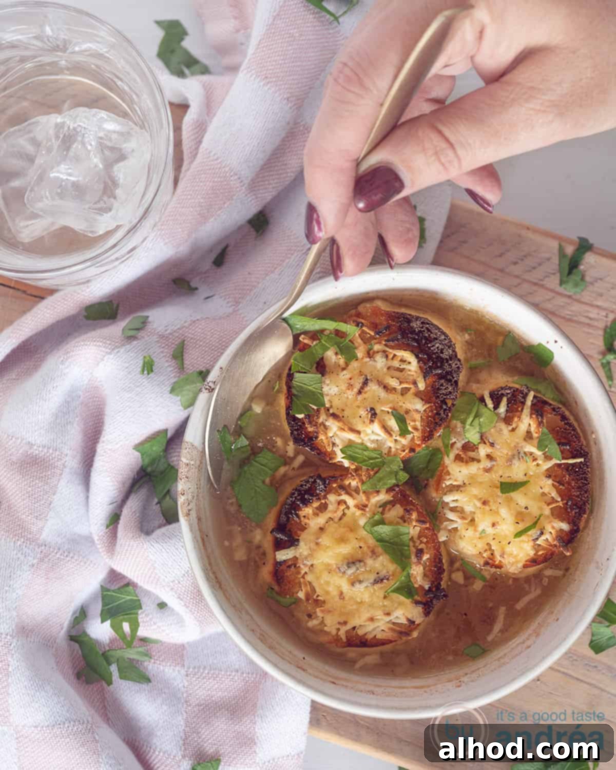 Simple Beer French Onion Soup 3 A white bowl with beer onion soup and three slices of gratinated baguettes in the soup. A hand with a spoon taking some soup. A pink white tea towel around the bowl