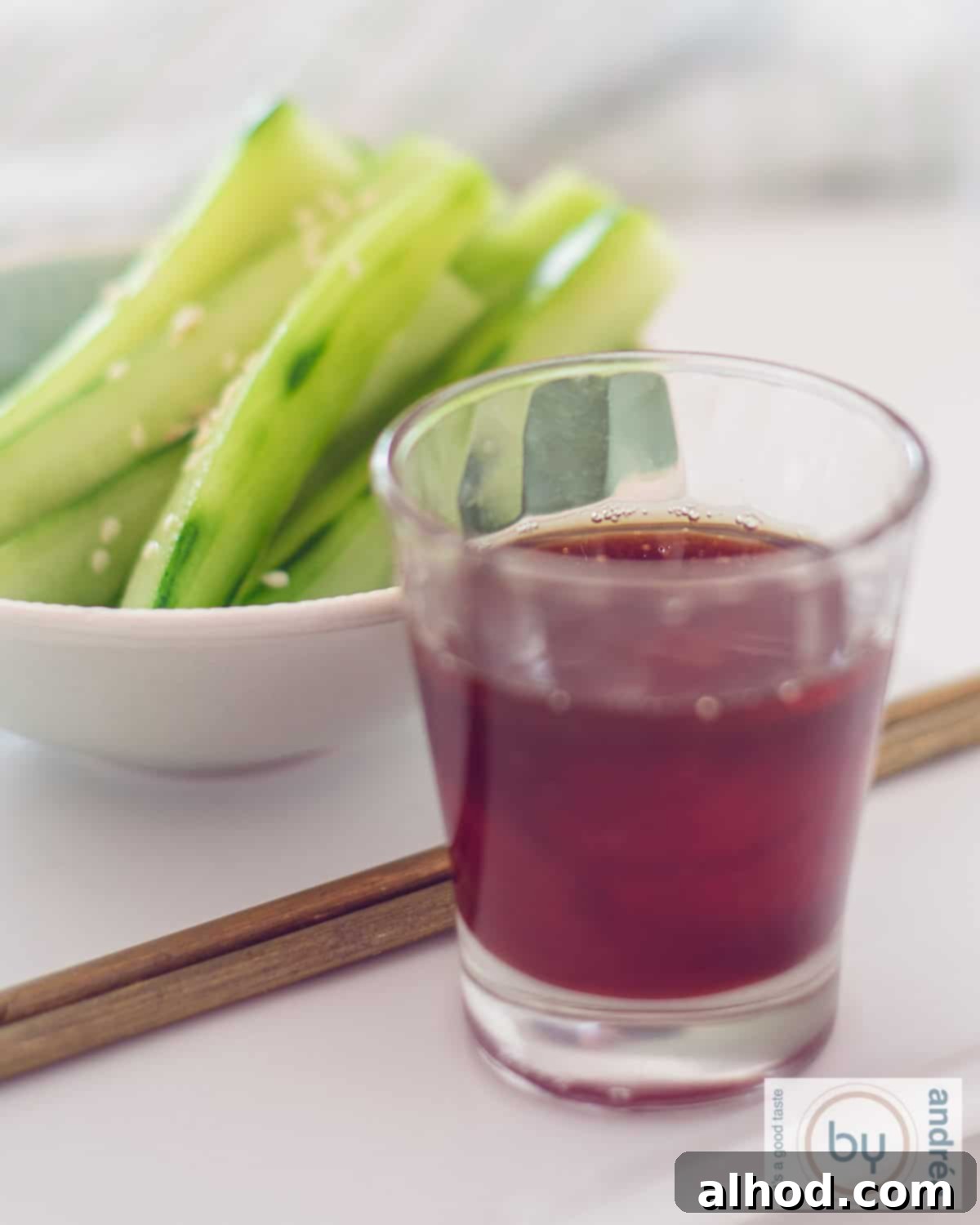 An aerial photo: in the foreground a glass with soy and chili sauce dip, then two chopsticks and in the background cucumber strips in a blue bowl.