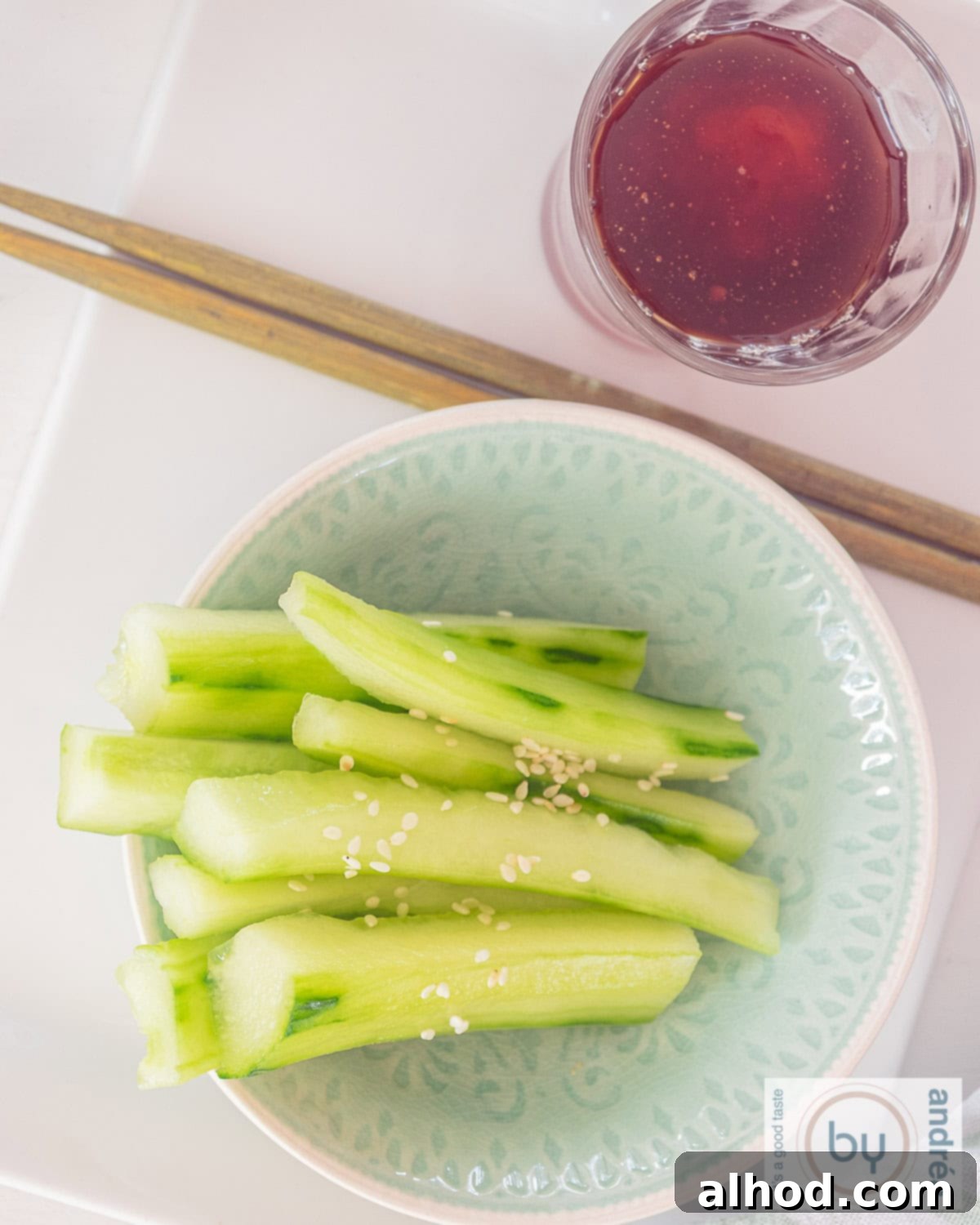 Spicy Sesame Cucumber Salad 3 A photo from above of a blue bowl with cucumber with sesame seeds and above that a glass with dip sauce. Two golden chopsticks lie in between.