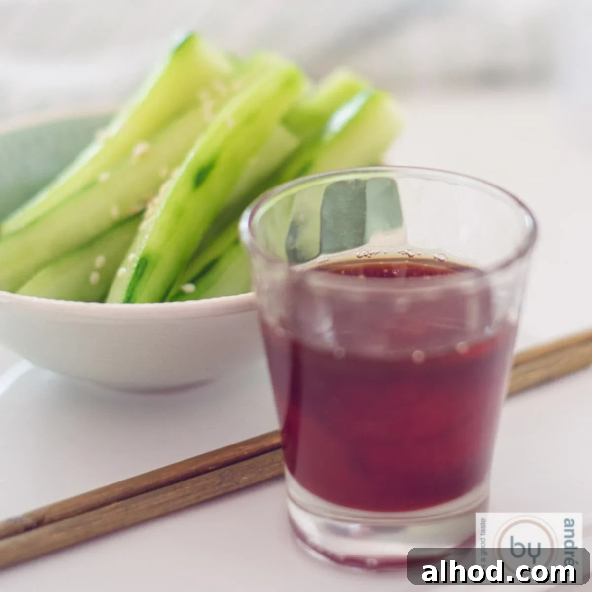 Spicy Sesame Cucumber Salad 4 A square photo with a bowl of cucumber and sesame strips in the background, two chopsticks and a glass with a spicy dipping sauce in the front.