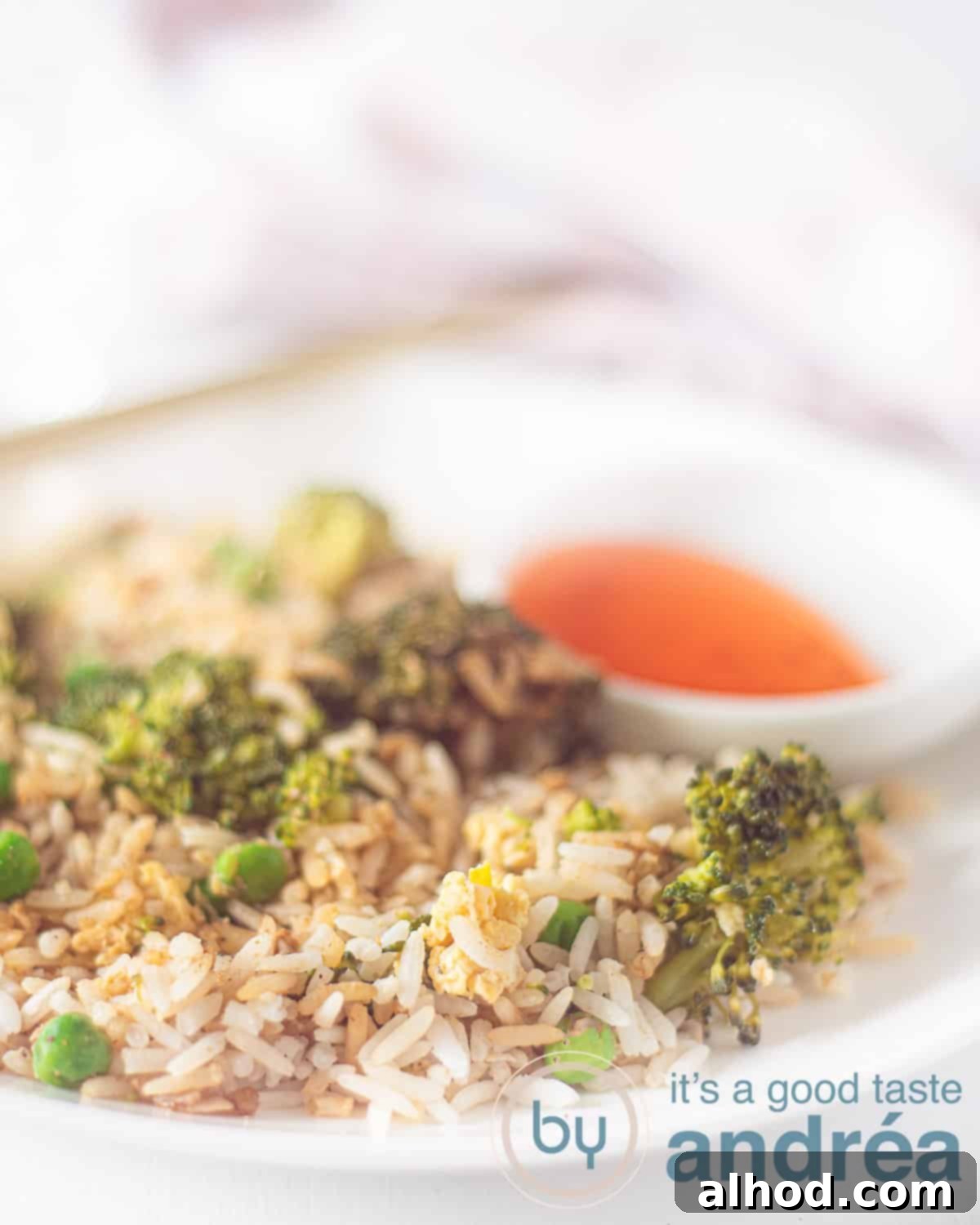 Velvety Egg and Vibrant Broccoli Fried Rice 2 A vibrant plate of Broccoli and Egg Fried Rice, featuring fluffy rice, green broccoli florets, scrambled egg, and sweet peas. A small bowl of sweet chili sauce sits beside it on a white background, inviting a delicious meal.