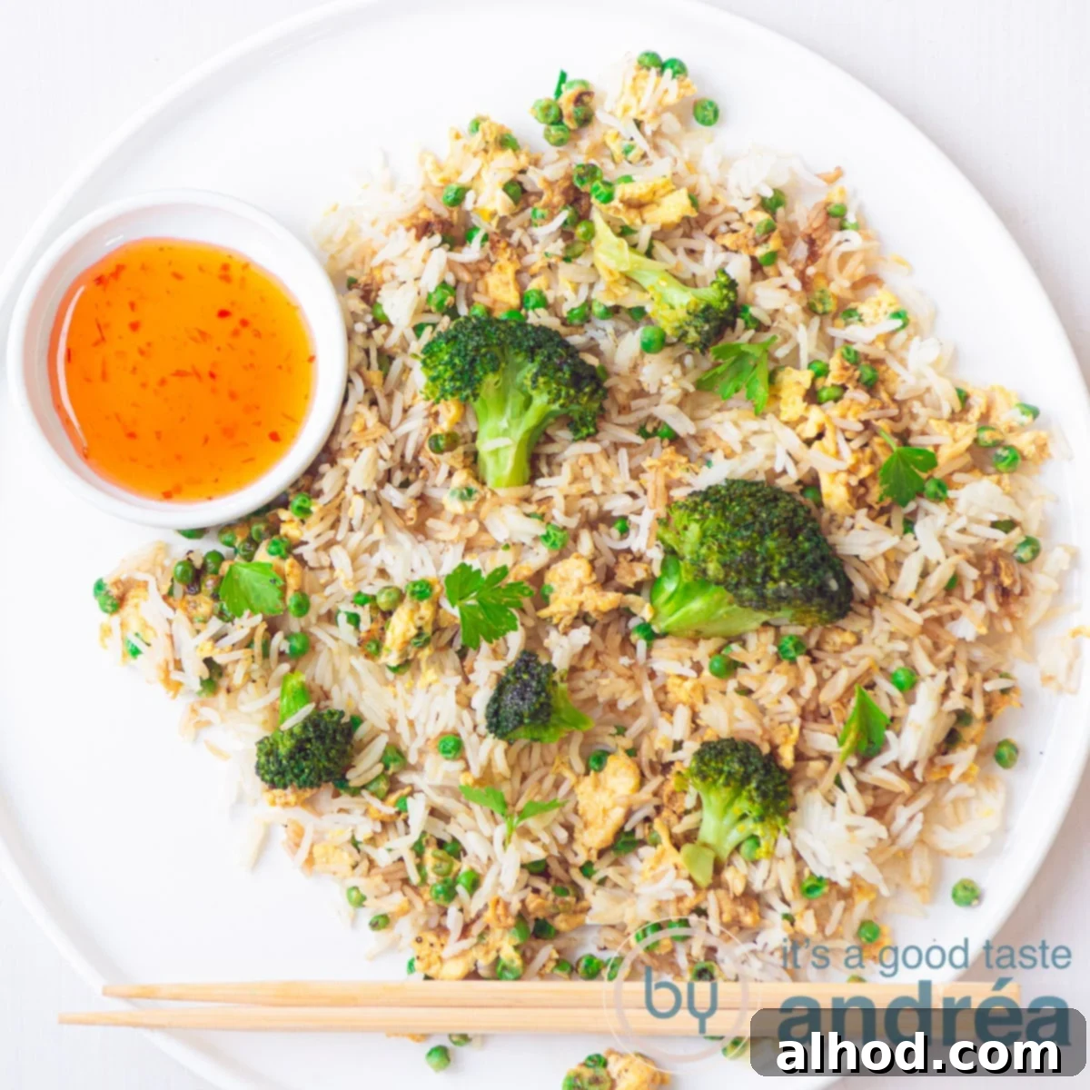 Velvety Egg and Vibrant Broccoli Fried Rice 6 A square photo of a white plate with fluffy fried rice, tender broccoli florets, sweet peas, and savory egg strips. A small bowl of chili sauce and two chopsticks complete the appetizing presentation.