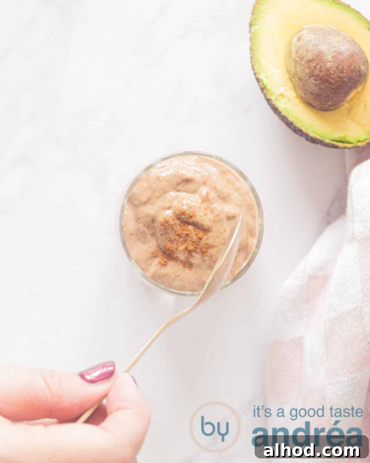 Silky Smooth Chocolate Avocado Treat 2 A photo from above of the Avocado smoothie with chocolate without banana. On a white background. On the right side a piece of avocado and a pink and white tea towel. A hand takes a scoop with a golden spoon