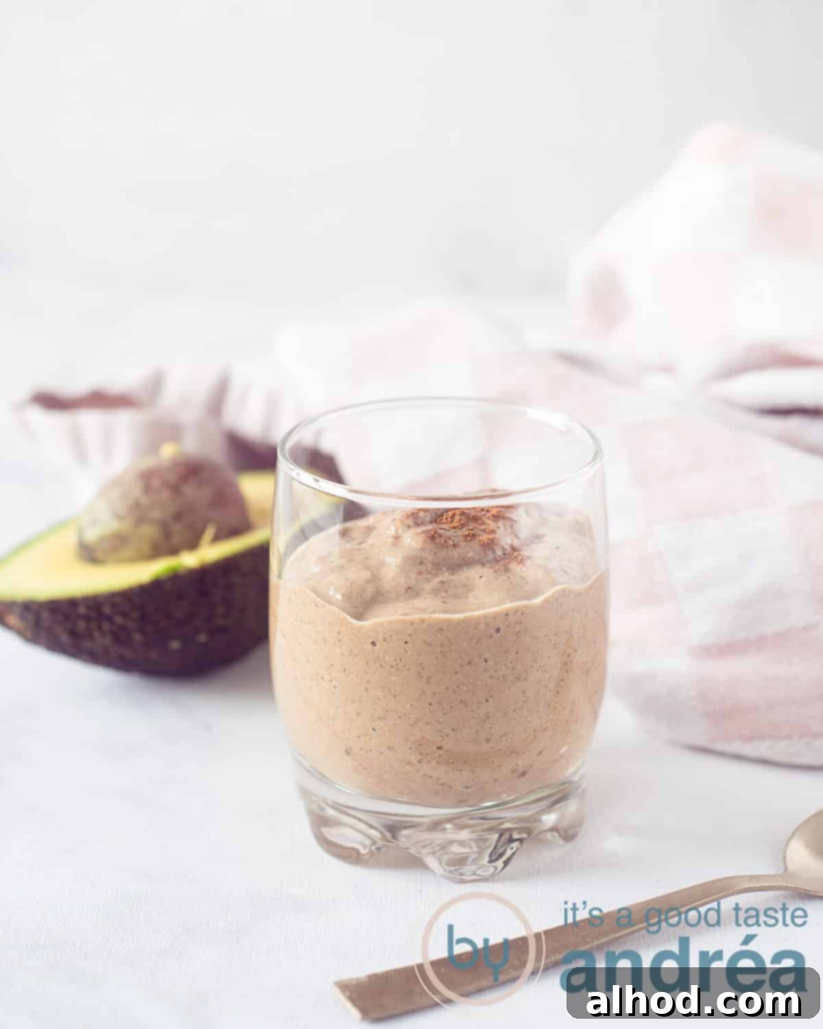 Silky Smooth Chocolate Avocado Treat 5 A glass filled with chocolate smoothie with avocado and coconut. A white background. In the back half a avocado and a white and pink cloth.