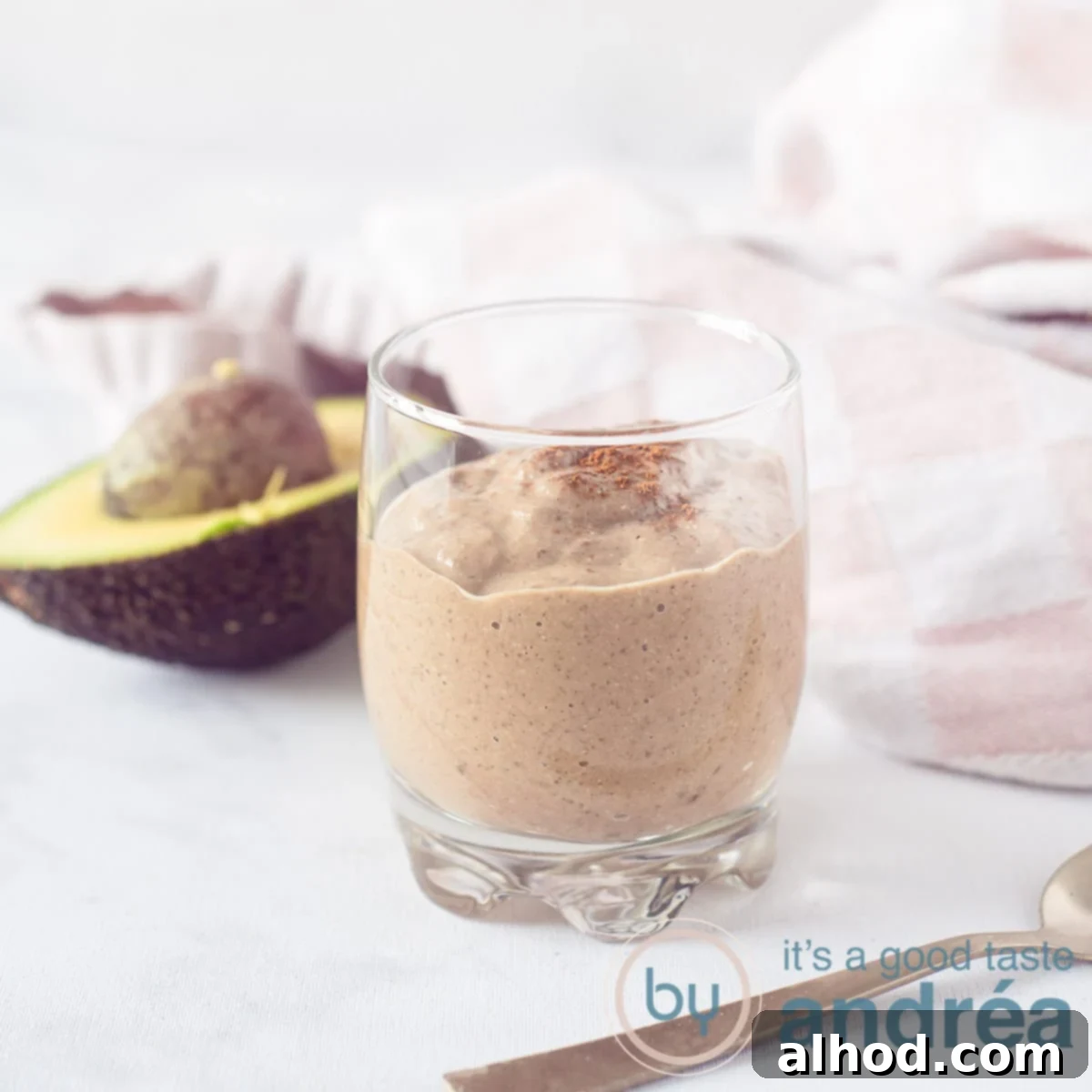 Silky Smooth Chocolate Avocado Treat 6 A square photo with a glass of chocolate coconut smoothie. An avocado and a pink white tea towel in the background.
