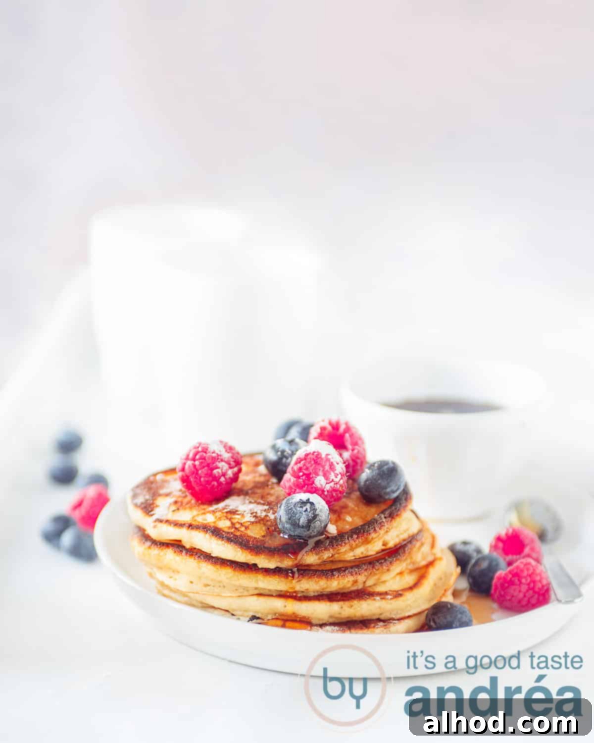 Ultimate Fluffy American Pancakes 2 A height photo of a white plate with American pancakes, fresh fruit. In the background a bowl with maple syrup and two white cups.