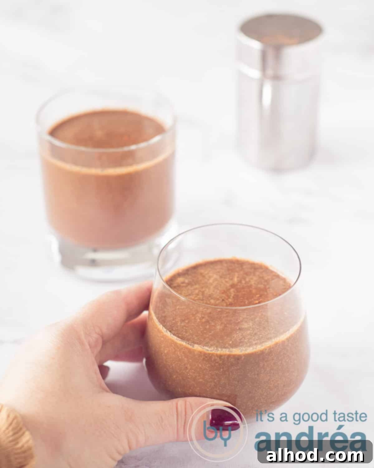 Cocoa Coconut Water Refresher 2 A height shot with two glasses filled with chocolate smoothie with coconut water, spinach and cherries. In the back right a cocoa spreader. A hand takes the front glass. A white background.