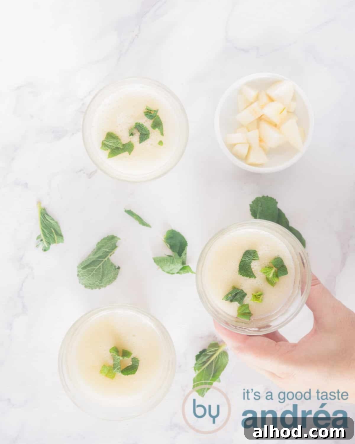A photo from above of three glasses filled with a pear, vanilla and coconut water smoothie with fresh mint. This is also on the white background around it. A hand takes a smoothie.