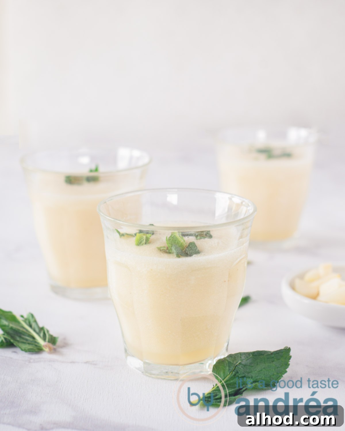 Three glasses filled with a pear, vanilla and coconut water smoothie on a white background. Some mint leaves scattered around.