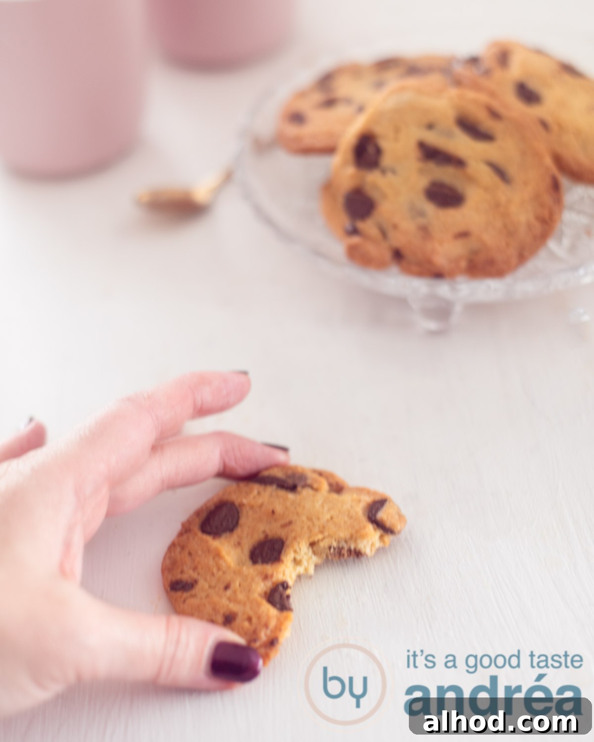 Your Personal Small Batch Chocolate Chip Treat 5 A white background. In the bottom a chocolate chip cookie with a bite. A hand holding it. In the back a glass plate filled with cookies.