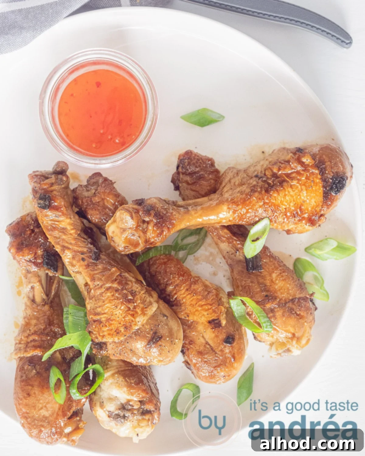 A photo from above of six chicken drumsticks bathed in a glossy soy sauce glaze, garnished with vibrant green spring onions. A small bowl of chili sauce sits to the side, ready to add an optional kick.