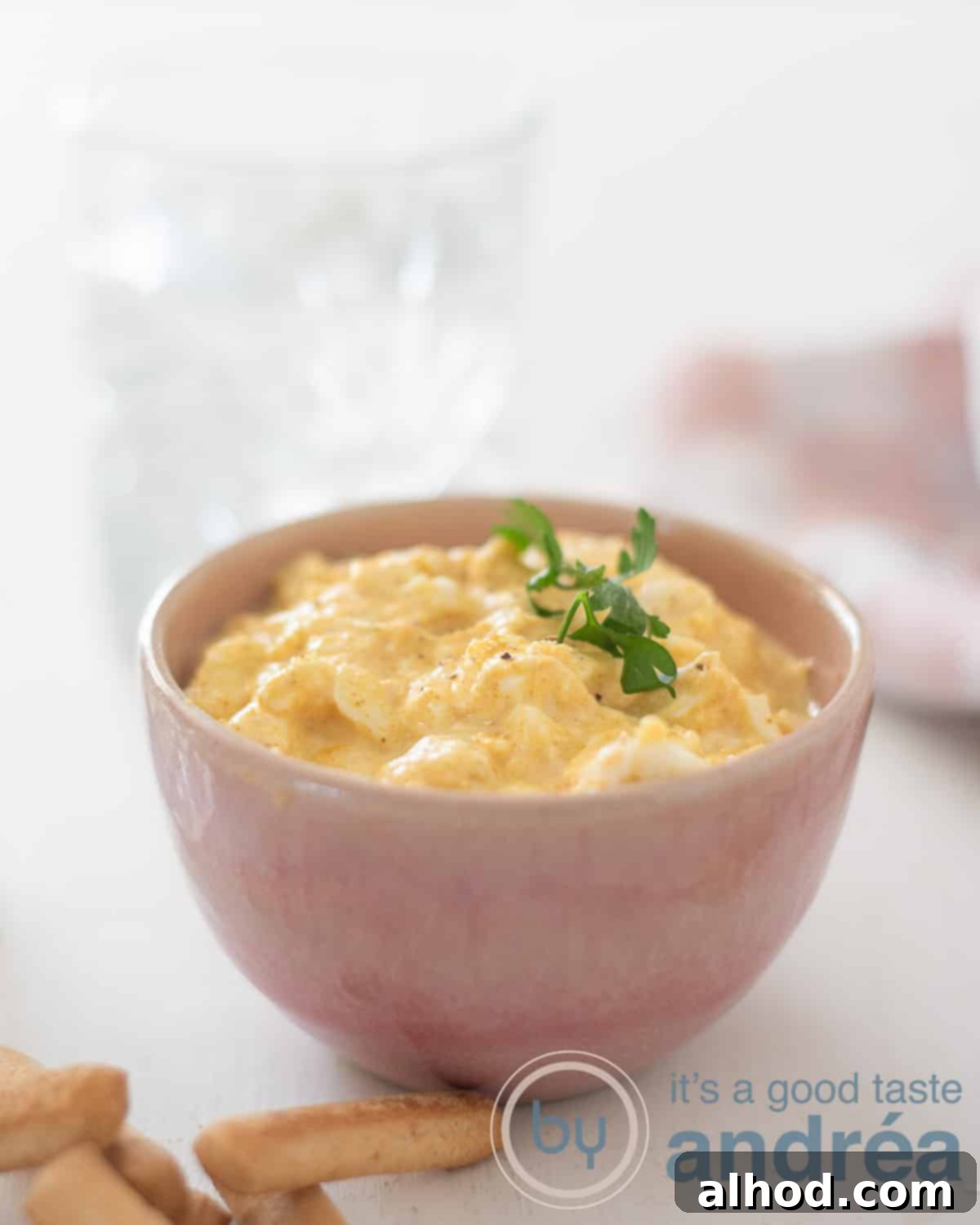 Reinvented Egg Salad No Mayo 2 A vibrant pink bowl filled with creamy egg salad, set against a clean white background. In the foreground, a few tapas breadsticks are ready for dipping, while a glass of water and a pink and white tea towel are subtly placed in the background, creating an inviting and fresh presentation.