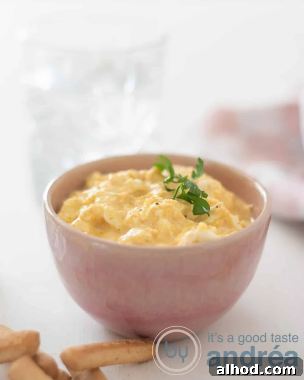 Fresh & Zesty Egg Salad 2 An elevation photo with a pink bowl filled with creamy egg salad in the middle on a white background. In the front are some tapas bread sticks. In the back a glass with water and a pink white tea towel.