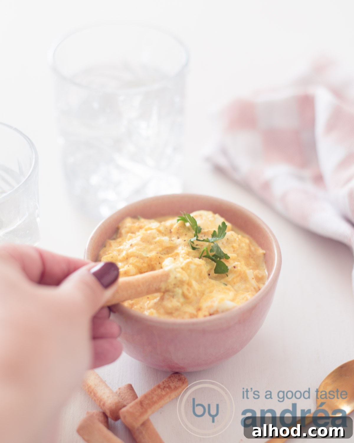 Reinvented Egg Salad No Mayo 5 A close-up, elevated photo of a pink bowl brimming with creamy egg salad, made without mayonnaise or mustard. A tapas breadstick is playfully dipping into the rich spread. The bowl rests on a pristine white background, highlighting the freshness and appeal of the dish.