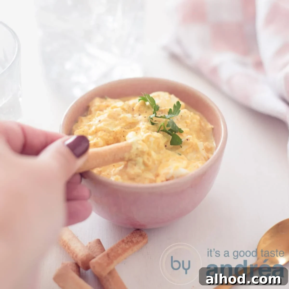 Reinvented Egg Salad No Mayo 6 A square photo of a pink container filled with an egg salad without mayonnaise. A tapas stick scoops something out. A white background. Top right a pink white tea towel