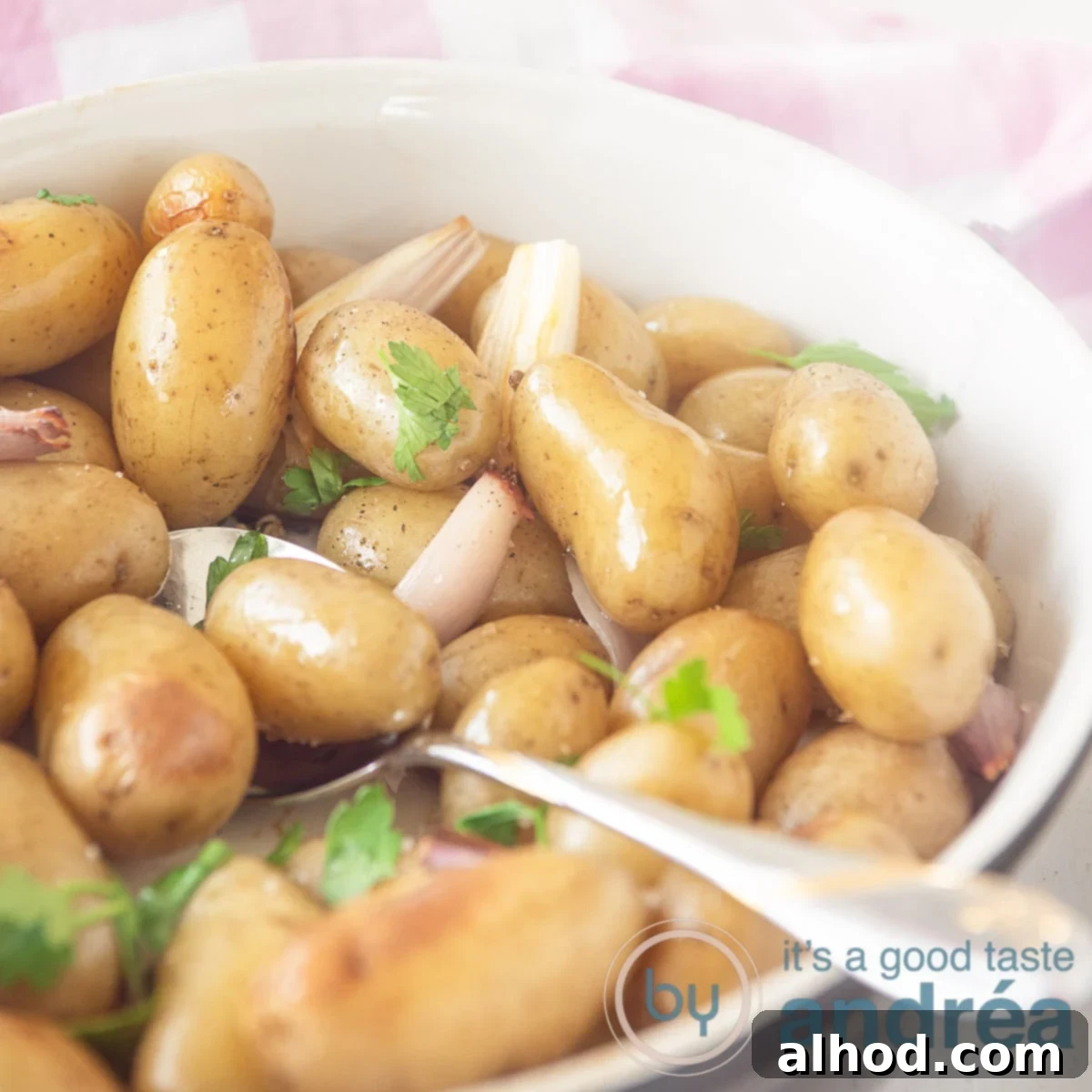Crispy Oven Roasted Baby Dutch Potatoes 6 A square photo with a black and white oven dish filled with potatoes with skin, shallots and herbs. A spoon sticks into the bowl.