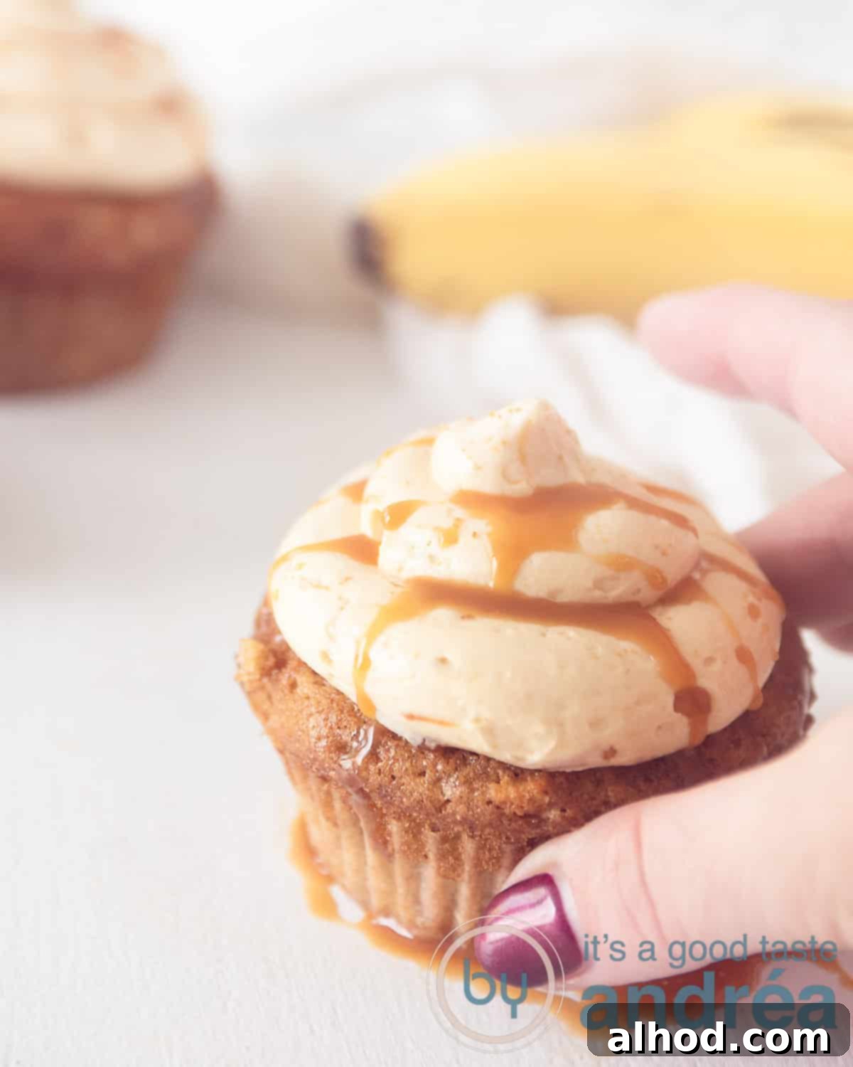 A hand reaches for a perfectly frosted banana cupcake with caramel buttercream, with more cupcakes and fresh bananas in the background, creating an inviting scene.