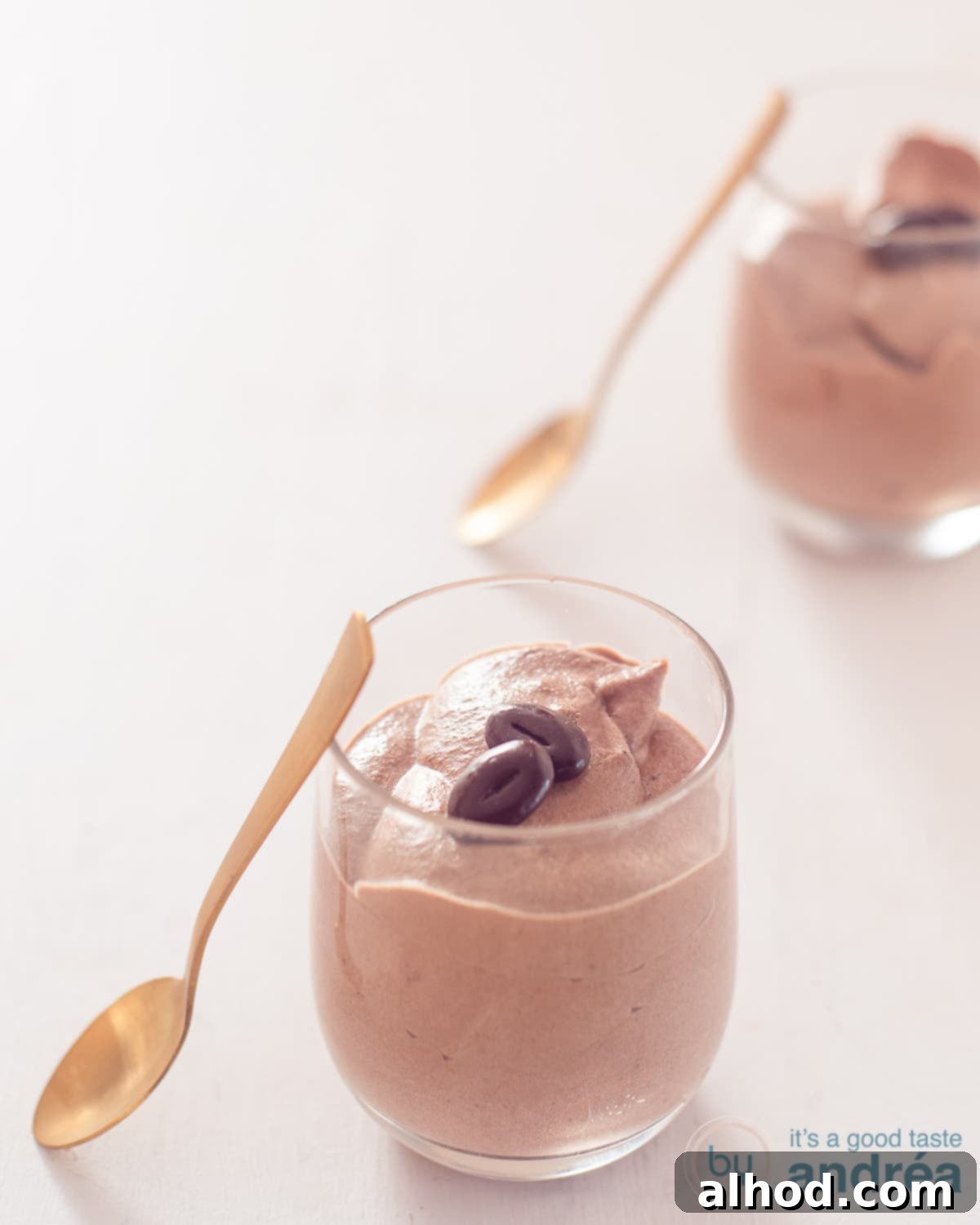 Velvety Milk Chocolate Mousse in Minutes 2 A height photo with two glasses filled with milk chocolate mousse. A gold spoon rests against the glasses. A white background.
