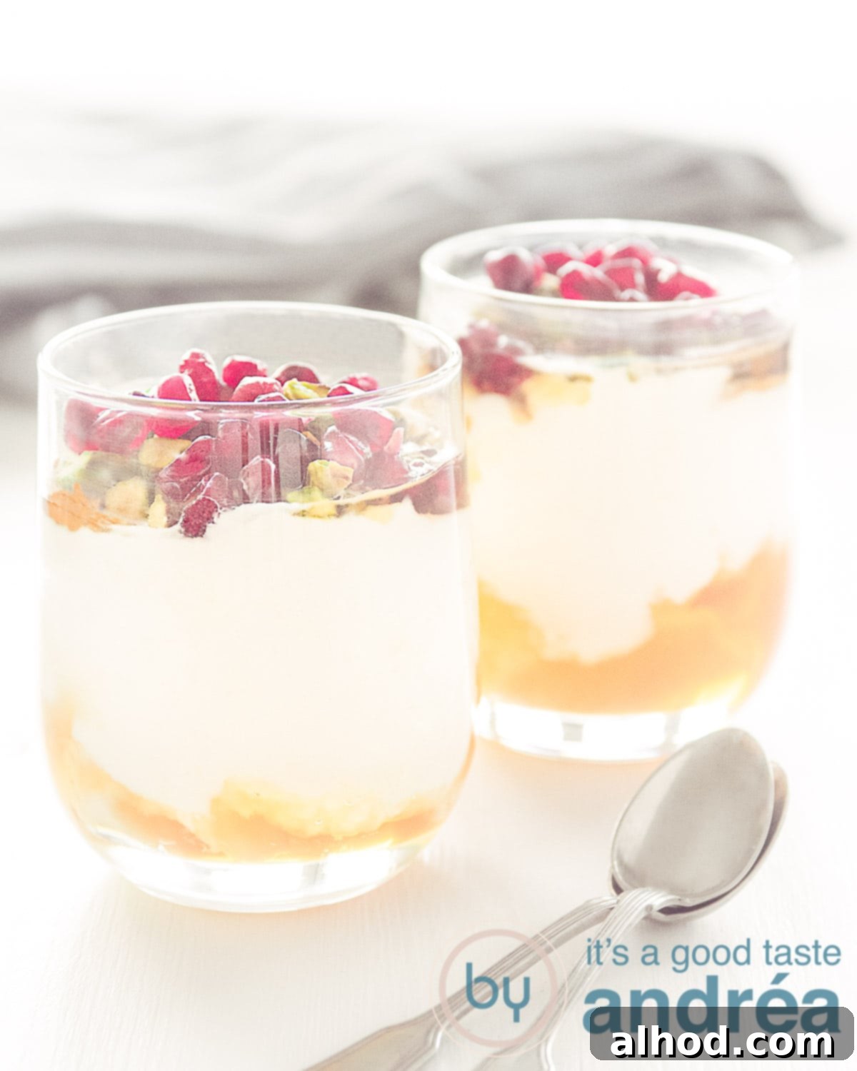 Two elegant glasses of yogurt with honey and pomegranate, garnished with loose pomegranate seeds and chopped pistachios on a light background.