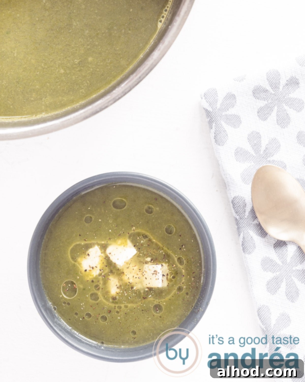 Hearty Vegan Spinach Potato Soup 2 A top-down view of a steaming bowl of creamy vegan spinach potato soup, garnished with fresh spinach leaves and a drizzle of olive oil. A golden spoon rests beside it on a white background, with a grey and white tea towel and a part of the soup pot visible in the frame.
