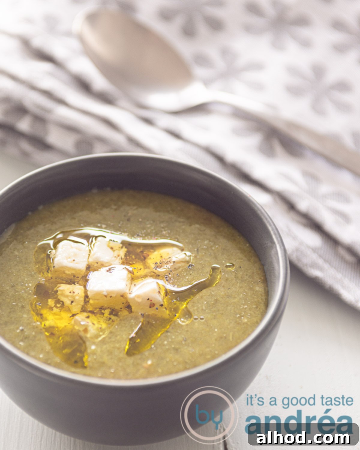 Hearty Vegan Spinach Potato Soup 5 A close-up shot of a bowl of creamy green spinach potato soup, beautifully presented in a grey bowl on a crisp white background. A grey and white patterned tea towel is partially visible in the top left, adding a rustic touch.