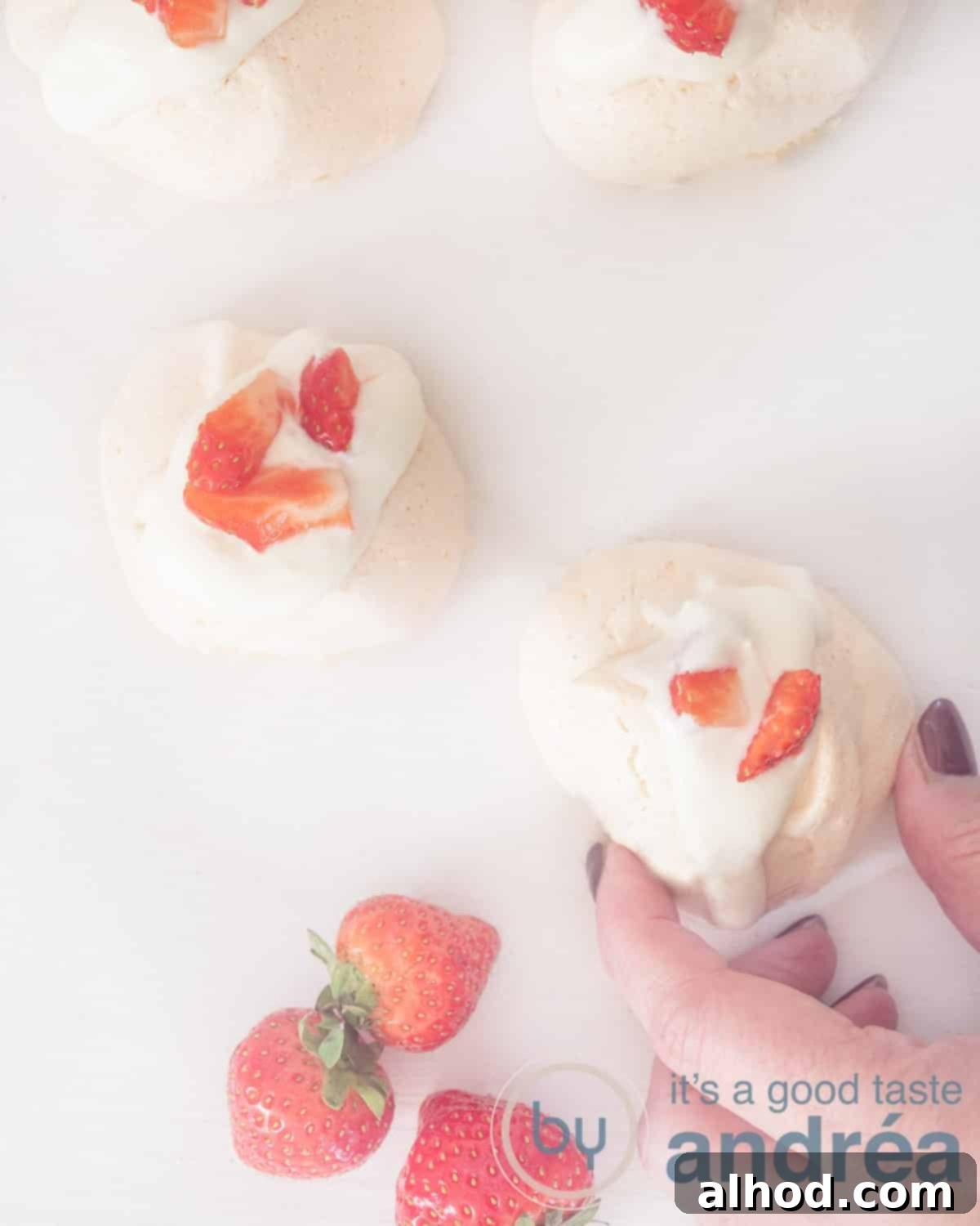Fragrant Rose Meringue Kisses 2 A photo from above of meringues with strawberries, four pieces. At the bottom right, a hand takes a meringue. Three strawberries are at the bottom left. The meringues are light pink, garnished with fresh rose petals, and arranged artfully with vibrant red strawberries.