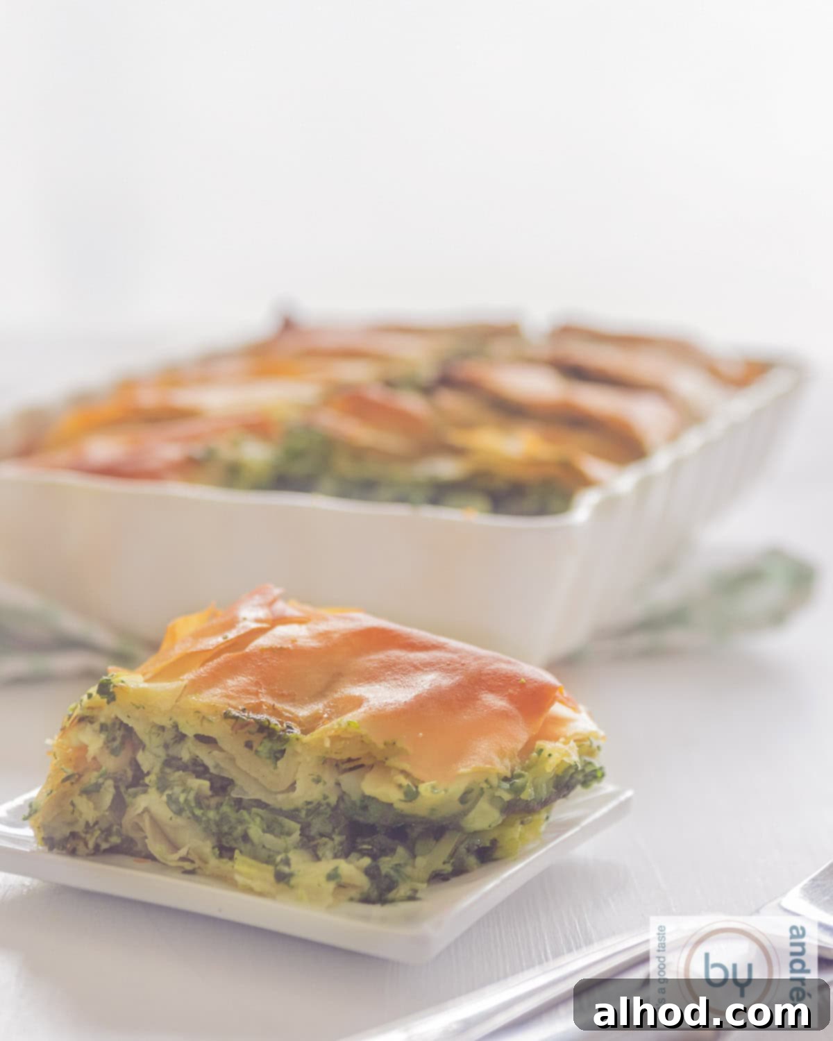 Simple Greek Spanakopita Pie 2 A photo of a plate with a piece of spanakopita. A casserole dish with several pieces in the background.