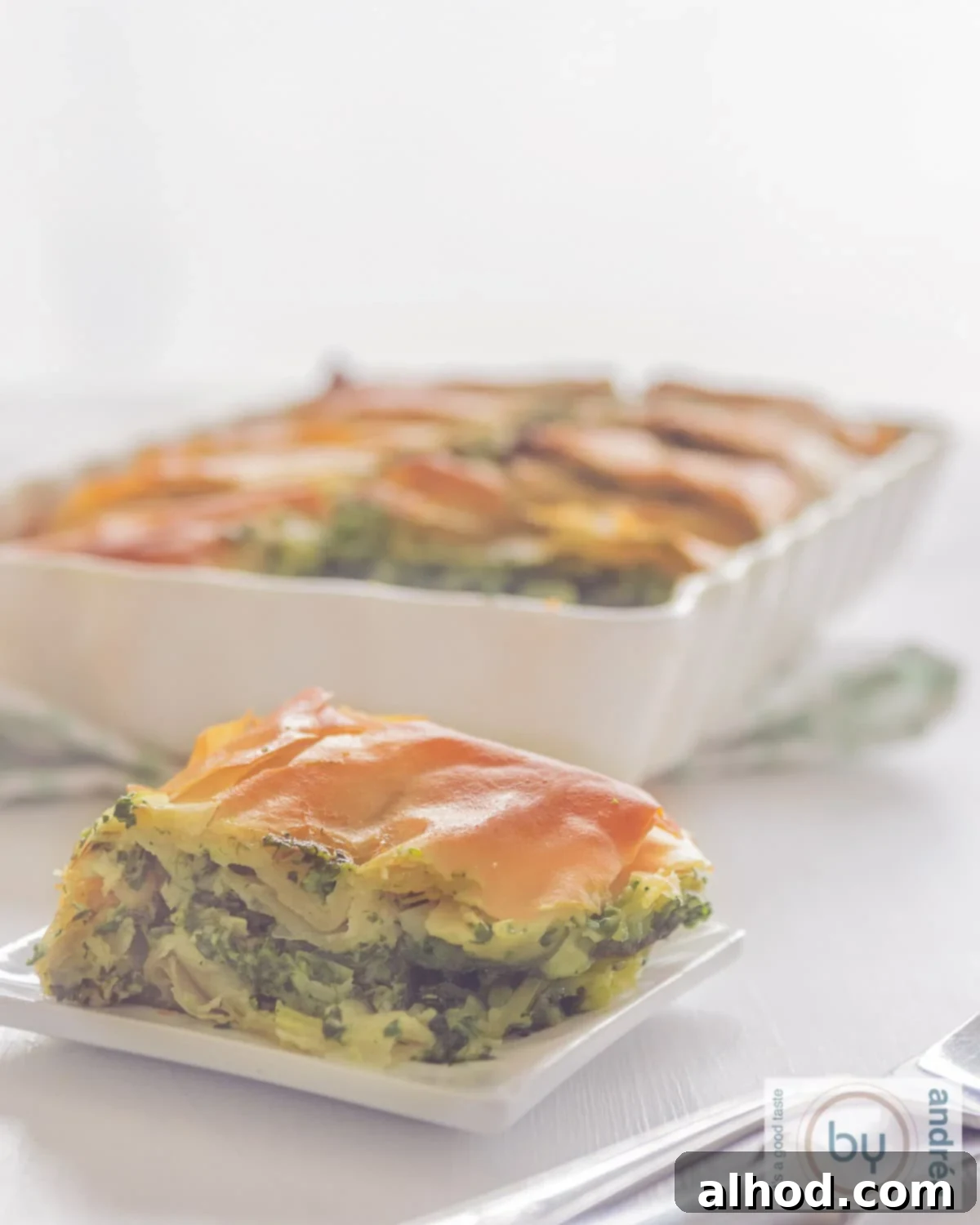 A photo of a plate with a piece of spanakopita. A casserole dish with several pieces in the background.