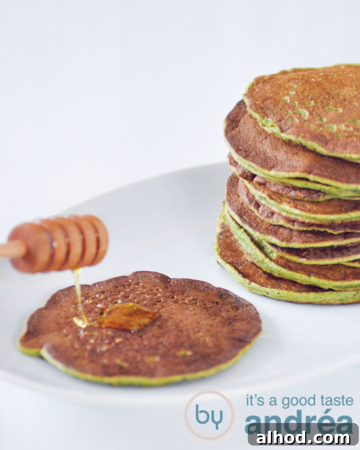 A beautifully arranged white plate featuring a stack of green spinach pancakes. One pancake at the top is artfully drizzled with golden honey, showcasing the deliciousness of this healthy breakfast option.