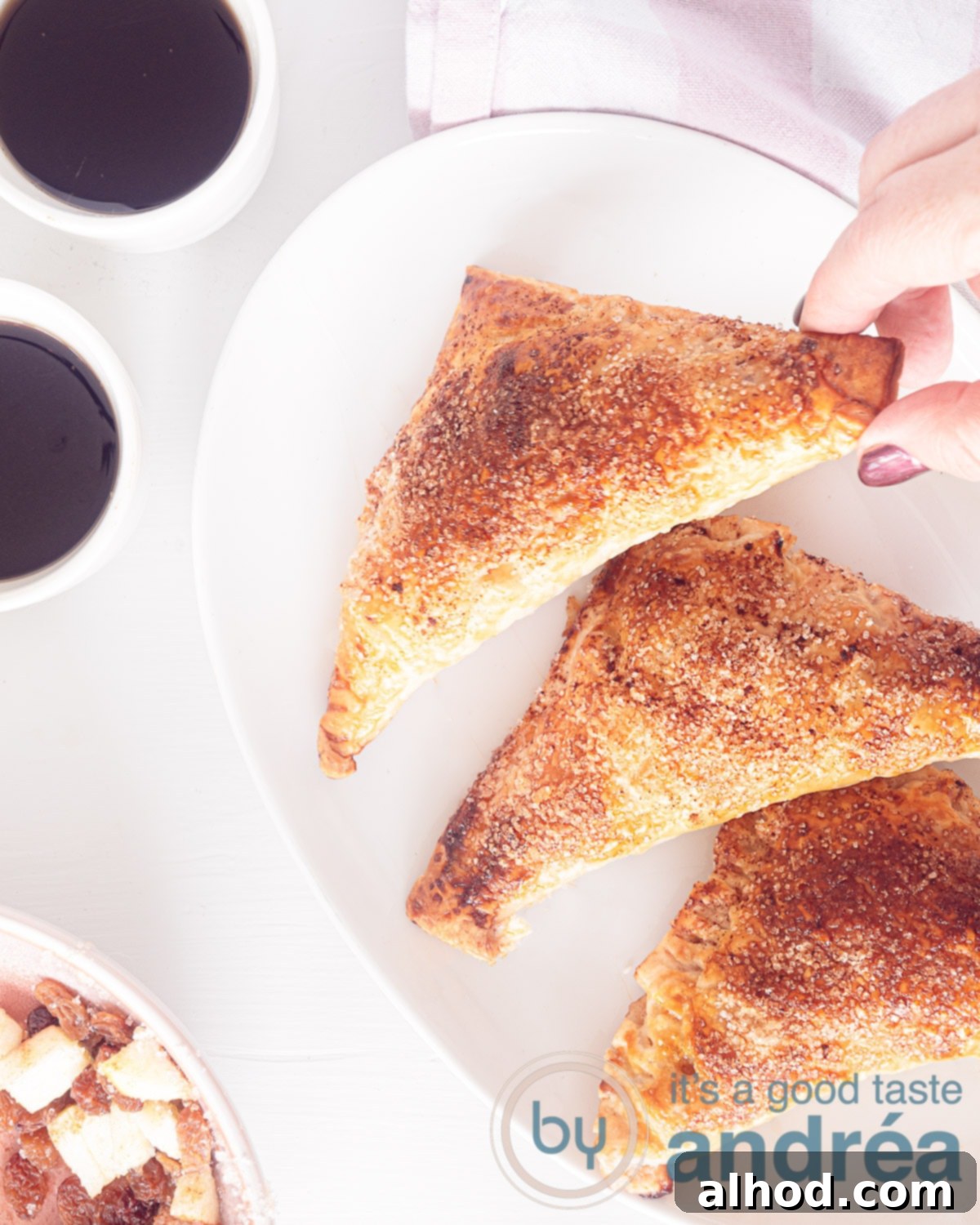 Classic Dutch Apple Turnovers 2 A photo taken from above of a white bowl with three apple turnovers, two white mugs with coffee on a white background, highlighting a delicious breakfast or snack scene.