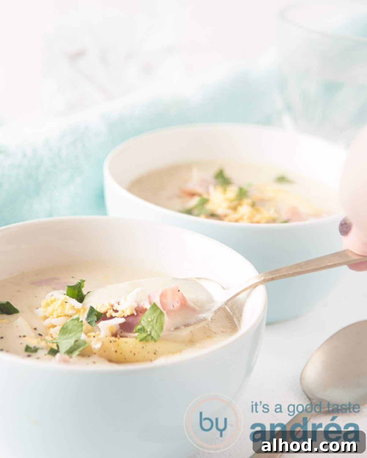 Velvety Dutch White Asparagus Soup 8 A white bowl with creamy Asparagus soup, garnished with ham strips and grated egg, with a spoon taking a spoonful, emphasizing its enticing texture.