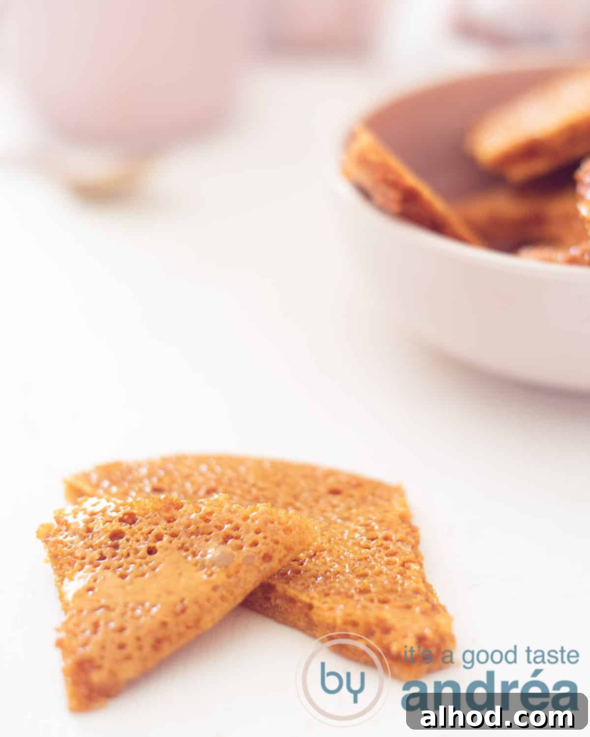 Two pieces of honeycomb toffee in the front on a white background. In the back right a pink bowl with various pieces.
