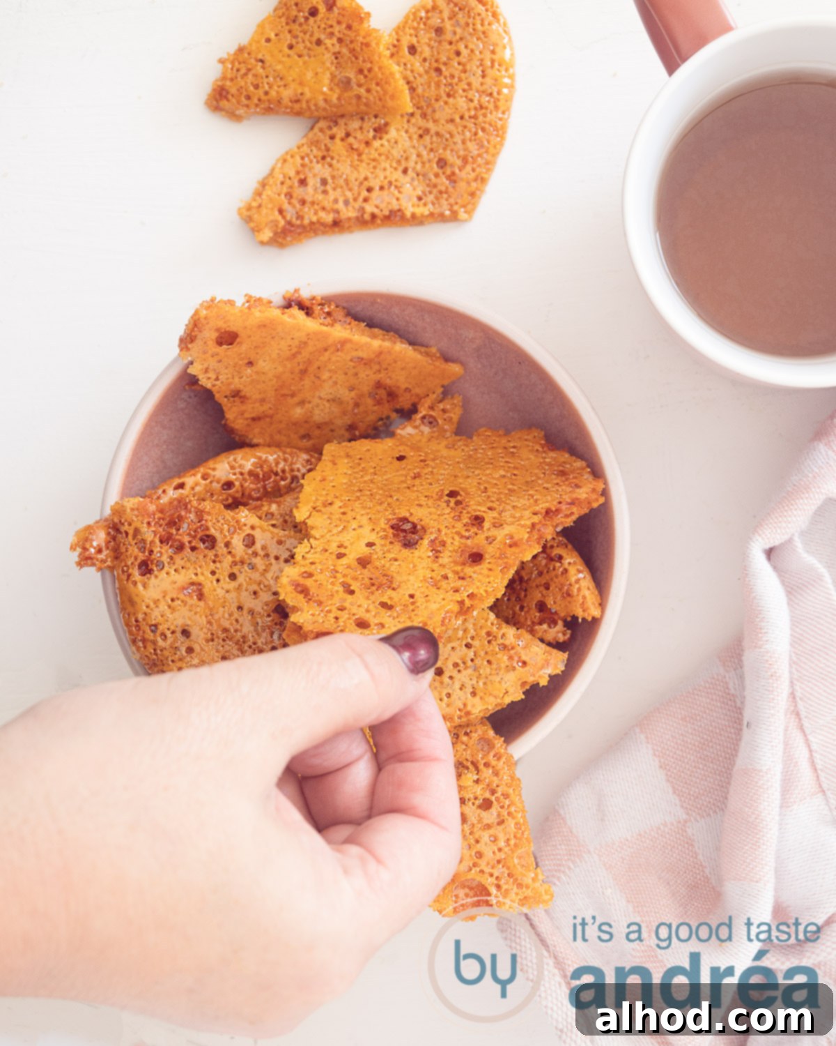 A hand takes a piece of honey comb from a pink dish. That is on a white background. Bottom left a pink white tea towel. Top right a pink cup with tea.