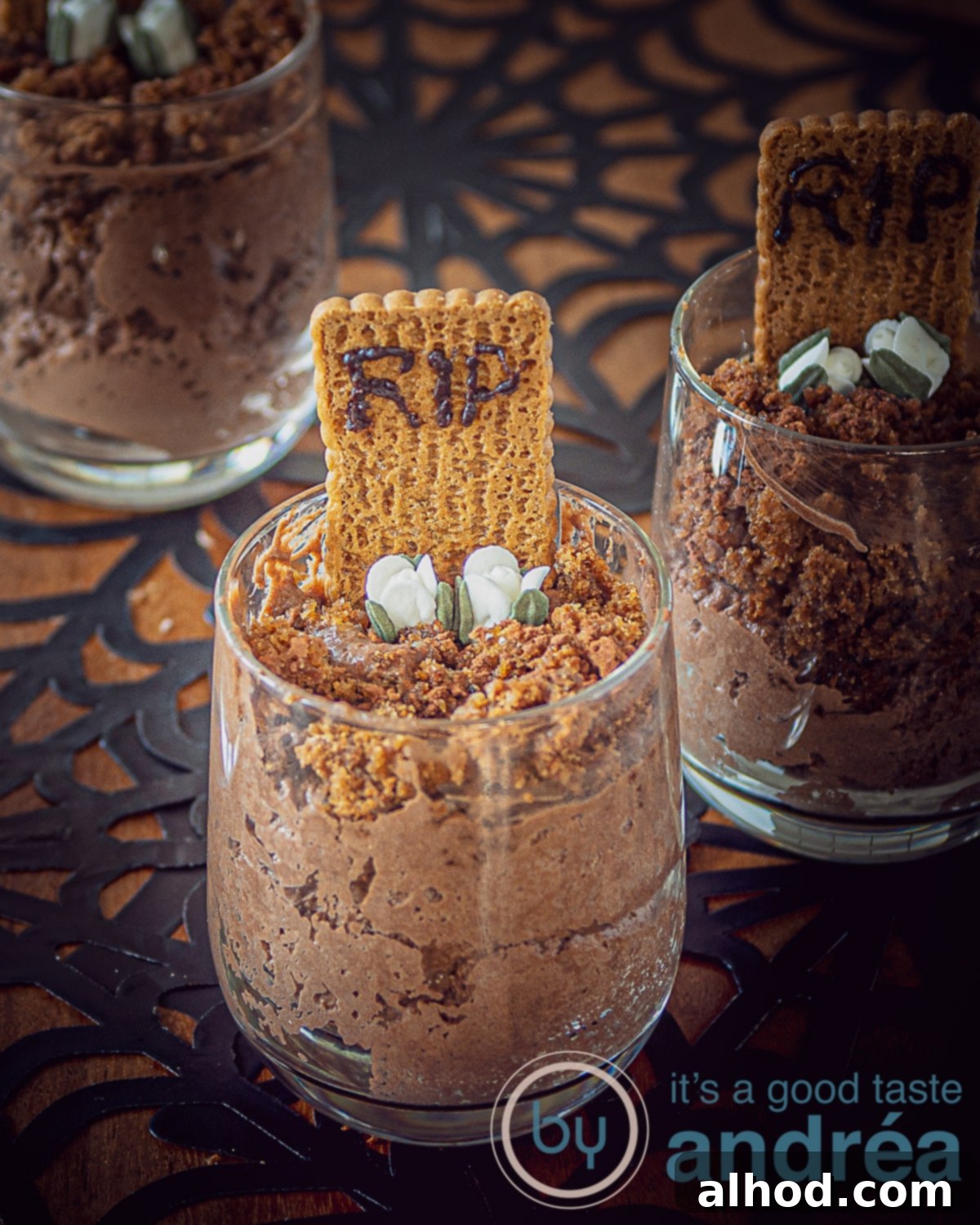 Easy Chocolate Graveyard Mousse 2 A large glass filled with brownies, chocolate mousse, speculoos biscuit with RIP and sugar flowers in front. Two more desserts on the right side and at the back left in the shadow