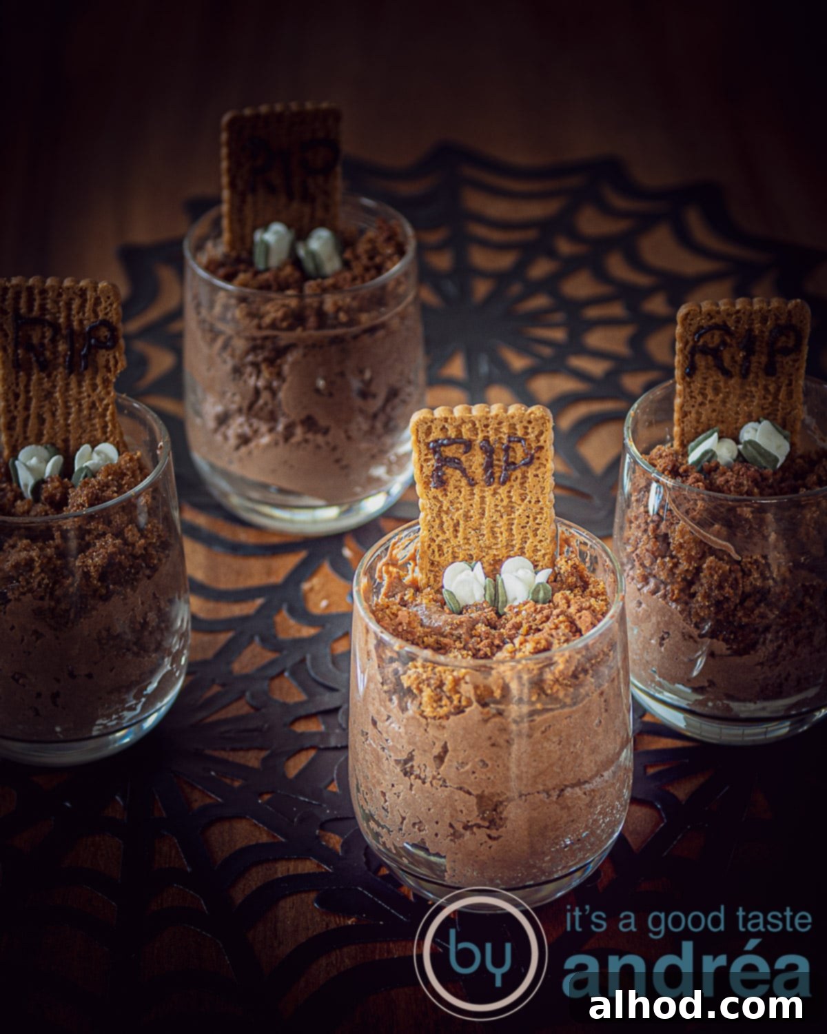 Easy Chocolate Graveyard Mousse 3 An elevation photo of a spotlighted glass with chocolate mousse, sugar flowers and a caramelized biscuit with RIP in it on a spider web background. Three more glasses in the shade around it