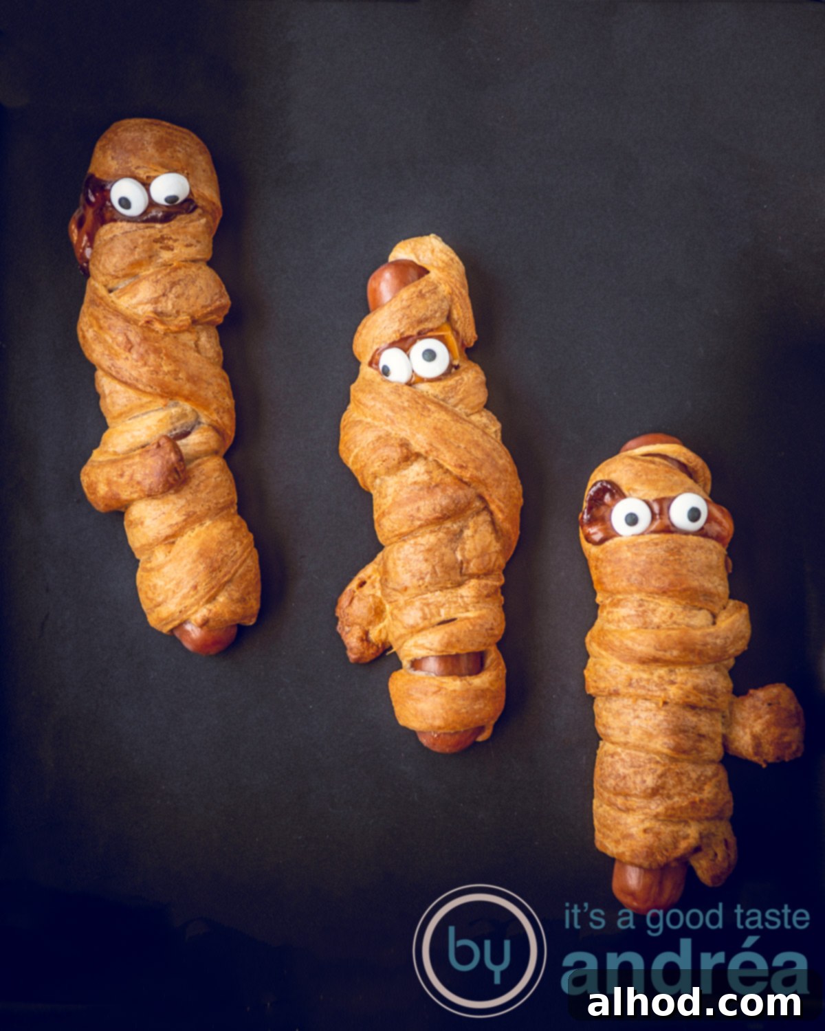 Halloween Hot Dog Mummies 3 Three golden-brown hot dog mummies with candy eyes on a black background, ready to be served for Halloween.