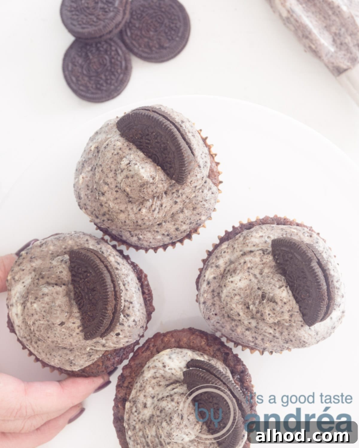 Simple and Scrumptious Oreo Chocolate Cupcakes 2 A photo from above of four Chocolate cupcakes with Oreo Frosting. A hand takes one. a white background. Top left a stack of cookies.