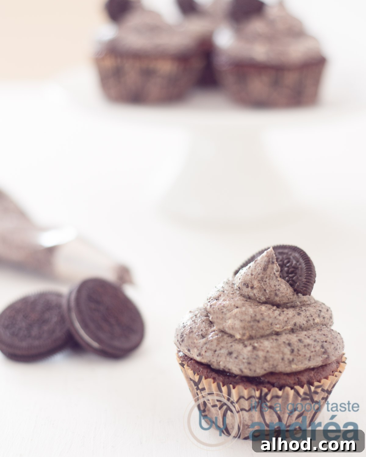 Simple and Scrumptious Oreo Chocolate Cupcakes 6 In front right an Oreo Cupcake on a white background. Left Oreo cookies and a piping bag. In the back a cake stand with chocolate oreo cupcakes
