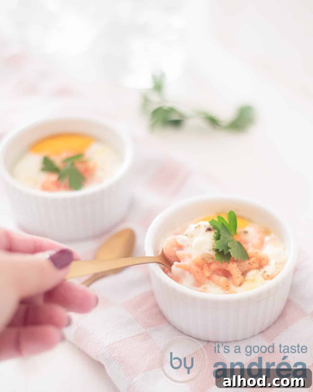 Silky Steam-Cooked Eggs in Ramekins 2 Two ramekins filled with oeuf en cocotte and shrimps. A hand takes a bite, showcasing the creamy texture. The dishes are elegantly placed on a pink and white tea towel.