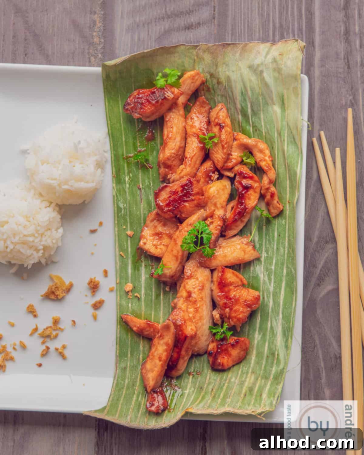 Whip Up Thai Chicken in 15 Minutes 3 A square plate, gray background. On the left are rice balls, on the right a banana leaf with four-flavored Thai chicken.