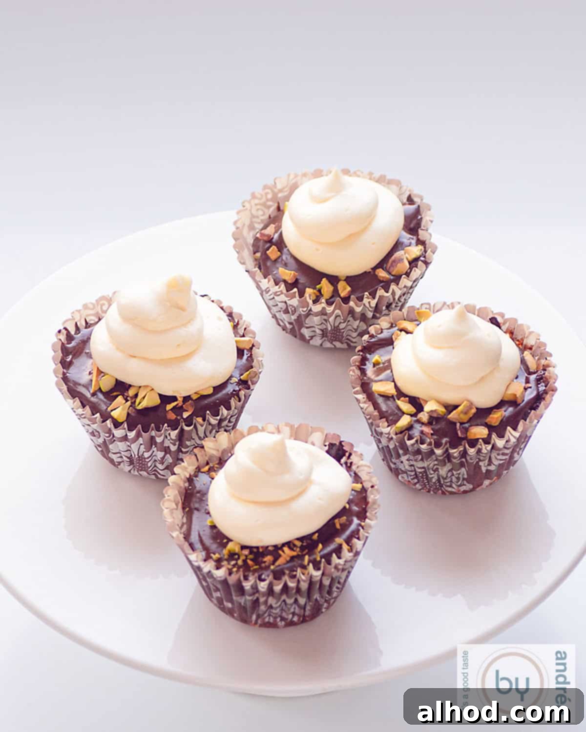 Decadent Triple Chocolate Cupcakes 2 A beautiful display of four Triple Chocolate Cupcakes on a white cake stand, adorned with dark chocolate ganache, white chocolate buttercream, and sprinkled with salted pistachios.