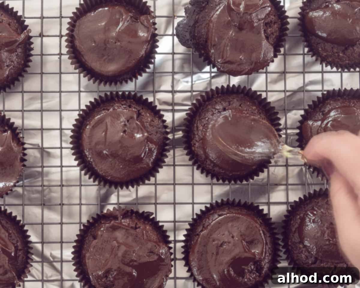 Decadent Triple Chocolate Cupcakes 6 Cooled chocolate cupcakes arranged on a wire rack, with a spoon spreading glossy dark chocolate ganache over one of them, preparing for the next layer.